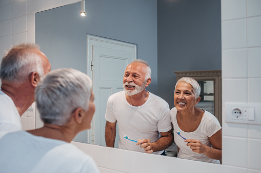 Elderly couple smiling and brushing their teeth in the bathroom mirror from Hoffman Dental in Hillsboro, OR