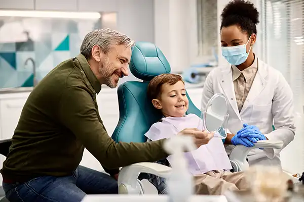 Father holding a mirror for his happy, young son sitting in a dental chair during his dental exam at Hoffman Dental in Hillsboro, OR