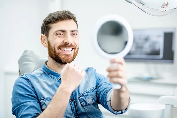 Happy patient checking out his handsome smile in a mirror while sitting in a dental chair