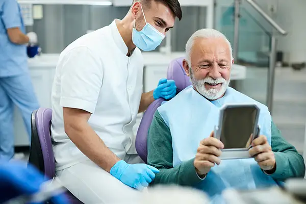 Happy, senior patient looking at himself in a mirror with a pleased male dental assistant.