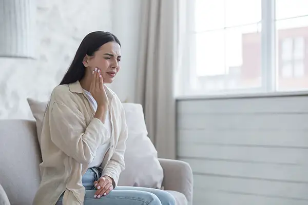 Young woman sitting on a couch, holding her cheek and wincing due to broken tooth, requiring emergency dental care at Jillian Prather Family Dentistry in Owasso, OK